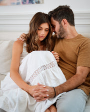 a man hugging and comforting his sad wife or partner while holding her hand on a white bed.